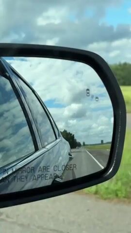 This video shows a rearview mirror from a car driving down a highway. The mirror reflects the sky with clouds, a road, trees, and a field. In the mirror, there are symbols indicating that other cars are present. The text "OBJECTS IN MIRROR ARE CLOSER THAN THEY APPEAR" is visible on the mirror.