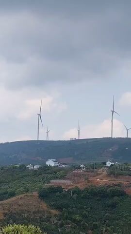The video shows a view of a hilly landscape with a small village in the foreground and several wind turbines on a ridge in the background.  The landscape appears to be covered in vegetation.  The sky is overcast with clouds. The wind turbines are rotating. There are a few houses visible within the village. One area appears to be undergoing construction with a wooden frame structure.