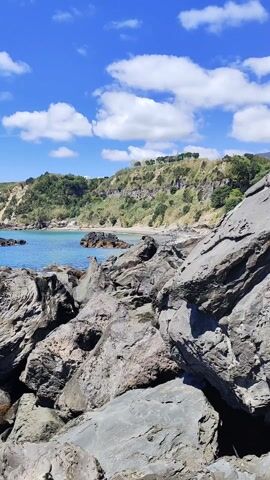 The video shows a rocky coastline on a sunny day. Large gray rocks occupy the foreground, transitioning to a view of a calm, turquoise sea. The background features a green, hilly coastline under a bright blue sky with scattered white clouds. There are no people visible in the scene. The camera pans slightly to the left, revealing more of the coastline and sea.