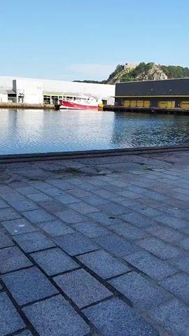 A vertical video shows a body of water with a red and white boat docked on the left side. Behind the boat are industrial buildings, one white and one with yellow doors. On a hill in the background, there are ruins. The foreground is a stone-paved walkway. The sky is clear and blue.