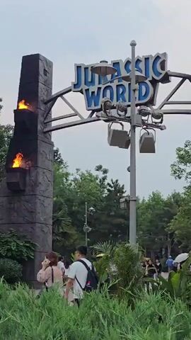 The media file shows an entrance to a Jurassic World-themed park. Dominating the frame is a large, gray, stone-like pillar with flames flickering from two openings, resembling a torch. Above and behind the pillar, a large sign with the words "JURASSIC WORLD" in blue lettering is visible, supported by a metal structure.

In the foreground, lush green bushes partially obscure the view. Several people are seen walking through the area. Closest to the camera, a woman in a light-colored dress with a black purse walks away from the camera. To her right, a man in a white t-shirt and black backpack walks in the same direction. Further back, more people are visible walking, some with umbrellas, suggesting it might be a warm or potentially rainy day. In the background, trees and a distinctive geodesic dome-like structure, reminiscent of a visitor center or attraction, can be seen.

The lighting suggests it is daytime, possibly late afternoon or early evening given the overcast sky. The overall atmosphere appears to be that of a popular tourist attraction. The setting is outdoors, in what appears to be a well-maintained park or theme park. The visible text on the sign clearly identifies the location as Jurassic World. The context provided states this is in Tongzhou District, China.