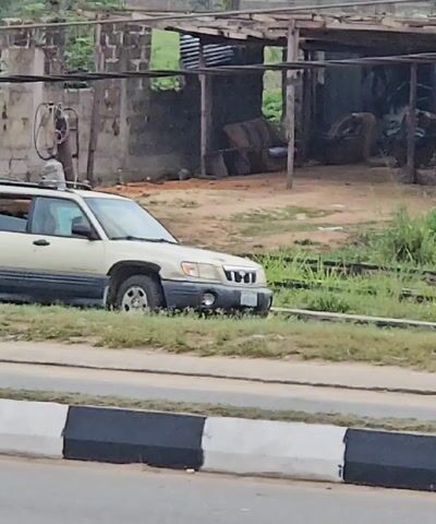 The video shows two people walking along the side of a road. One person is carrying a small orange basket. They are both wearing casual clothing and sandals. A beige SUV is parked on the other side of the road. Behind the SUV is a partially dilapidated building with furniture inside. The road has a black and white curb. The area appears to be a residential or semi-rural area with some vegetation.