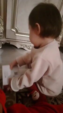 A toddler sits on a patterned rug in front of a light-colored, ornate cabinet. The toddler wears a light-colored long-sleeved shirt and red pants with a Santa Claus face on the knee. They push a small box across the floor towards the cabinet. A person wearing red lies on the rug at the edge of the frame.