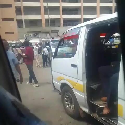 The video shows a scene from inside a vehicle, looking out at a bustling street scene.  Focus is drawn to a white van with its passenger-side door open, revealing a man seated inside wearing brown shoes. A few people walk by the van; including one man wearing a light blue shirt and another man in a green shirt. The backdrop consists of an aged building with many windows. The overall atmosphere conveys a busy and commonplace setting in what is probably an urban environment. The camera view appears to be taken from within another vehicle, with the car's interior visible on the left side.
