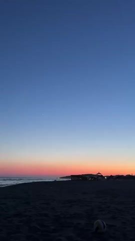 The video shows a beach at dusk. The sky transitions from a deep blue at the top to orange and pink hues near the horizon. The sea is visible on the left, with gentle waves. The beach is dark sand, and in the foreground, a volleyball rests on the sand. In the background, silhouetted structures, possibly buildings or beach huts, are visible along the coastline. There are no people or animals in the frame. The scene is calm and quiet, bathed in the soft light of sunset.