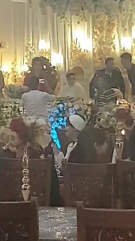 The video shows a gathering, possibly a wedding or celebration, with several people present. In the foreground, a table with floral decorations is visible. In the middle ground, a group of people are gathered, some sitting and some standing. One person is taking a photo with a phone. There are also two young boys in the middle ground, one dressed in a black shirt and white pants, the other in a patterned shirt and dark pants. In the background, a stage area is set up with a white backdrop and floral arrangements. Several other individuals are visible in the background, some in formal attire. The lighting appears to be dim and artificial, with colored lights illuminating the floral decorations. The overall environment seems to be an indoor venue with red carpeted flooring.