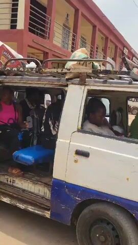 The video shows a white van with a partially open sliding door, exposing the interior. The lower portion of the van's side panel is blue and shows signs of rust. The camera moves to show a yellow and black three-wheeled vehicle, a TVS, positioned between the van and another similar blue and white vehicle. A person is visible inside the yellow vehicle. The vehicles are on a dusty, sandy-colored surface, likely a road or yard. A building with a reddish-brown exterior can be seen in the background. The scene suggests a transportation or transit environment.