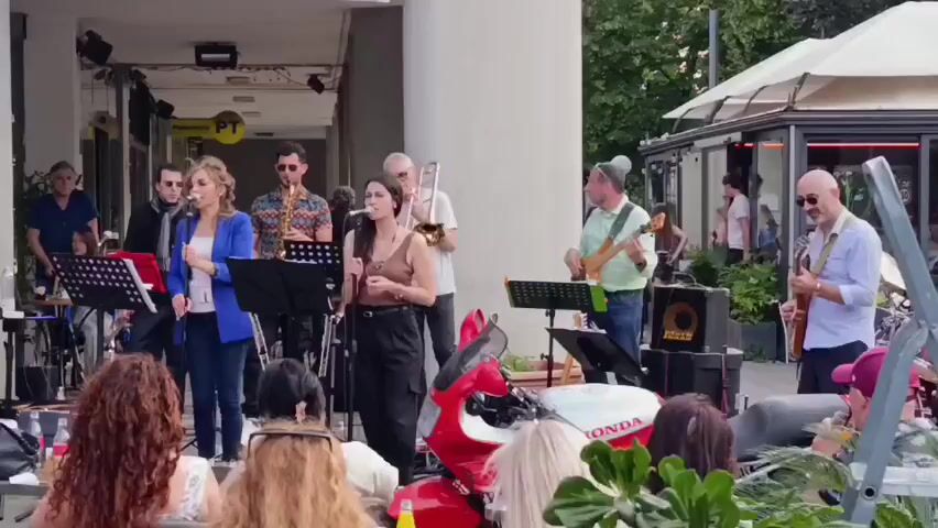 A band of eight musicians performs on an outdoor stage in front of a building. Two women sing into microphones. Other band members play a trumpet, saxophone, trombone, bass guitar, and electric guitar. Music stands with sheet music are visible. A red scooter sits in front of the stage. An audience sits and stands in front of the stage and outside a nearby cafe. The weather appears sunny.