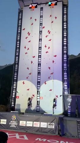 This video shows two individuals competing in a speed climbing event on a tall, two-lane artificial climbing wall. Each lane has red climbing holds arranged in a pattern up the wall. Digital timers are mounted at the top of the wall above each lane, displaying the current time. Projected circles of light illuminate the lower portion of each climbing lane where the competitors are positioned. Both competitors are wearing harnesses and are actively ascending the wall, using the hand and foot holds. Spectator banners and logos, including "WORLD CLIMBING" and "HBO max", are visible at the base of the structure. The surrounding environment appears to be outdoors, with a clear sky and mountains visible in the background. The competitors are in mid-climb, demonstrating speed and agility as they progress upwards.
