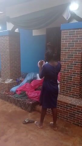 A woman wearing a blue dress blows up balloons outdoors in front of a decorated entryway. The entryway has two brick pillars, a light blue wall, and draped fabric above. There are several balloons in her hands after she blows them up. Her shoes are resting on the ground beside her feet. In the background, more balloons and draped fabric can be seen inside the entryway. There is a sound of music playing.