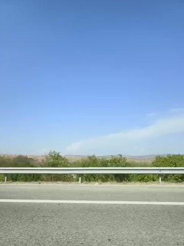 The video shows a view from a vehicle traveling along a paved road. A white metal guardrail runs parallel to the road, separating it from a grassy area with scattered bushes and trees. The sky is a clear, bright blue with a few wispy white clouds. In the distance, there appears to be a flat, open landscape, possibly farmland, with some vegetation. The camera angle suggests the video was recorded from the passenger side window of the vehicle. There are no people visible in the video. The environment appears to be rural or semi-rural.