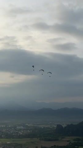 The video shows a landscape with mountains and a town in the distance. The sky is cloudy with patches of sunlight. In the sky, there are three paragliders flying. The paragliders are moving from left to right. One of the paragliders appears to be falling behind the other two.