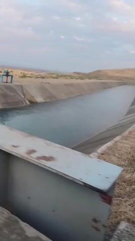 The video captures an outdoor scene, likely in a dry, arid environment with rolling hills in the background. The foreground shows a concrete structure, possibly a bridge or overpass, with a metal beam running horizontally across the bottom of the frame. Below this structure is a large, man-made canal or aqueduct filled with water. The canal is lined with concrete and appears to be quite wide, curving gently to the right and disappearing into the distance. The water in the canal is calm, with a slight ripple effect. Beyond the canal, the landscape transitions to a sandy, barren terrain with sparse vegetation. In the middle distance, a small blue structure or object is visible on the edge of the canal. The sky above is overcast with soft clouds. The camera is in motion, giving a sense of movement through the scene. The overall impression is one of a large-scale water management system in a desolate landscape.