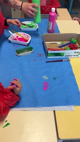 A group of people, likely children and an adult, are engaged in a painting activity at a table covered with a blue cloth. The adult's arm, tattooed and wearing a red sleeve, is visible reaching for a green bottle of paint. A child's arm, wearing a red smock, is also visible, with pink paint on their hand. Other objects on the table include bottles of green and pink paint, a red basket, a white tray with pink paint and a brush, a green brush, a cardboard box filled with art supplies like a paint roller and a toy vehicle, and a paintbrush. There are splatters of pink and green paint on the blue cloth, indicating active use. The environment appears to be a classroom or art studio setting.