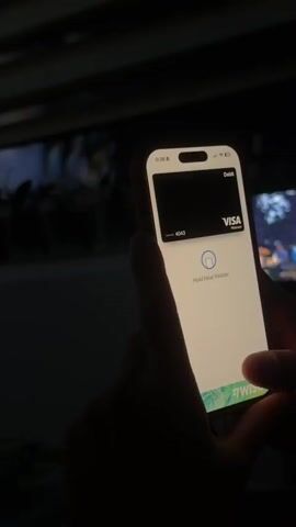 The video shows a close-up of a hand holding a smartphone with a credit card displayed on the screen. The phone is displaying an interface that indicates a payment is pending, with the text "Hold Near Reader." The background is dark and out of focus, with some indistinct shapes that might be furniture or a screen. The lighting suggests the scene is indoors and dimly lit. The video is very short, lasting only a few seconds.