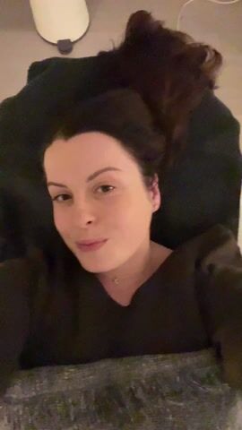 A young woman with dark hair and fair skin is lying down, looking directly at the camera. She has dark hair styled in a way that suggests it's pulled up or behind her head, but parts of it are visible spread out above her. She is wearing a dark-colored top, possibly brown or black, with a v-neckline, and a delicate gold-toned necklace with a small crescent moon charm. Her expression initially is a subtle, closed-mouth smile.

She is positioned on what appears to be a bed or a massage table, covered by a dark blanket or sheet, with another lighter-colored blanket or throw partially visible towards the bottom of the frame. Behind her head, to the left, there's a white cylindrical object, possibly a humidifier or an air diffuser, with a dark cap. The wall behind her is a light, neutral color, transitioning to a soft green hue towards the top right of the frame. As the video progresses, her expression shifts slightly, and she pouts briefly. The camera also moves slightly, revealing electrical outlets and a white cord plugged into one of them on the green-colored wall in the background. The lighting is soft and even, suggesting an indoor setting.