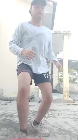 A person wearing a light blue long-sleeved shirt, black shorts, a black baseball cap, and grey athletic shoes is seen performing a dance routine in an outdoor courtyard. The person is facing right in the initial frame, with their arms slightly extended and their right leg raised. As the video progresses, the person moves their arms and legs in a rhythmic manner, transitioning into a pose where they are facing forward with their legs spread apart and their arms bent at the elbows. The background features a light-colored building with a doorway, a broom leaning against the wall, and several pairs of flip-flops scattered on the ground. The courtyard floor is made of concrete and appears worn and discolored. The sky is overcast, suggesting a cloudy day.