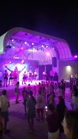 The video shows a nighttime outdoor event with a stage and an audience. The stage is lit with purple and blue lights, and there are several performers dancing on it. The audience, composed of adults and children, is gathered in front of the stage, also illuminated by the stage lights. Some people in the audience are dancing, while others are watching the performance. The environment appears to be an open area, possibly a park or a venue with a paved ground.