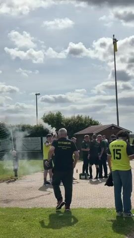 A wide, eye-level shot shows a crowd of people, mostly men, gathered outdoors in front of a small wooden building, a flag pole, and a chain-link fence. Many of the people are wearing yellow and black shirts, some of which say "SV KLINKUM" and some have numbers, like "15." In the foreground, on the left, a man in a yellow shirt holds a smoke flare that emits white smoke. Another man, wearing a black shirt that also says "SV KLINKUM," stands with his back to the camera, walking toward the group in the center. In the background, a man on the far right wears a yellow shirt with the number "15" on the back. The group of men in the center are gathered around a table or box, interacting with each other. The sky is partly cloudy. The ground is a mix of green grass and a paved area.