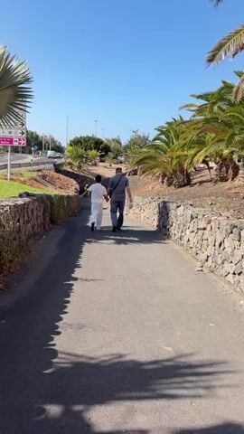 Two people, a man and a woman, walk away from the camera down a paved path. The woman is on the left, wearing a white outfit, and the man is on the right, wearing a grey shirt and dark pants. They appear to be holding hands. The path is bordered by low stone walls. On the left side of the path, there is a road with a few cars visible and a signpost with multiple directional arrows. Palm trees and other vegetation are present on both sides of the path, with dry earth and rocks visible beyond the walls. The sky is clear and blue, indicating a sunny day. The camera is positioned behind the couple, following them as they walk.