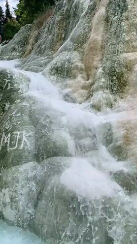 A close-up shot shows water cascading down a rocky, textured surface. The rocks appear to be mineral deposits, with streaks of green and white. Some faint white markings, possibly carved initials, are visible on the left side of the rock face. The water is white and foamy as it flows, creating a dynamic and textured effect against the static rock. The bottom of the frame shows a pool of light blue water where the cascade ends. The overall impression is of a natural, mineral-rich hot spring or waterfall.