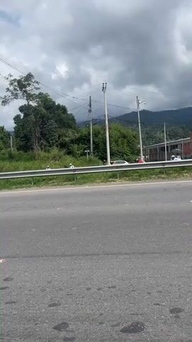 The video shows a road scene. On the left side of the road, there is an old, white building partially obscured by greenery. Adjacent to the road is a metal guardrail. Beyond the guardrail, several people are visible, seemingly gathered. A few motorcycles are parked nearby. In the background, utility poles with wires stretch across the sky, and hills are visible in the distance under a cloudy sky. The road surface is gray asphalt with scattered debris.