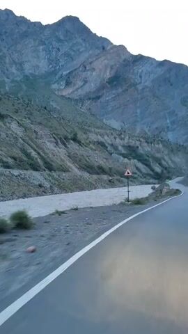 The video begins with a shot of a road alongside a mountain and a river. A warning sign depicting a downward arrow is visible on the right side of the road. The road curves to the right as it heads into a mountain pass. The sky is clear and the light suggests either early morning or late evening. The camera appears to be mounted on a vehicle, as evidenced by the motion blur and the perspective from the road. The mountains are rocky and barren, with some sparse vegetation on their slopes. The river is wide and appears to be flowing swiftly. As the video progresses, the road continues to wind through the mountains, with the river remaining on the left side. The landscape remains consistent, with the road leading deeper into the mountain valley. The video ends with a view of the road disappearing into the distance between the imposing mountain slopes.