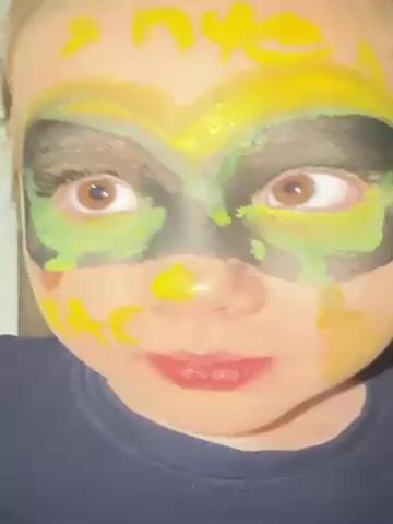 A young girl with light skin, brown eyes, and light brown hair is looking directly at the camera. She is wearing a dark blue shirt. Her face is covered in face paint. There is a mask-like design around her eyes in black and green. Yellow paint is also smeared around the mask and on her forehead. Her lips are slightly parted. The background is a blurry, light-colored wall.