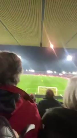 A video captures a spectator's perspective at a football (soccer) game, filmed from the stands. The angle is upward, showing the stadium's roof and the bright floodlights illuminating the field. The field is visible in the distance with the goalpost prominently situated in the center. A crowd of people is visible beyond the field, suggesting a sizable audience. The video is taken from the viewpoint of someone sitting behind a few people, their heads and backs partially obscuring the full view of the field. There is audible cheering, indicating excitement in the stadium.