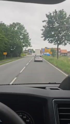 The video provides a first-person view from inside a vehicle, likely a car or van, driving on a two-lane asphalt road in Jade, Germany. The immediate foreground shows the vehicle's dashboard, including a portion of the steering wheel and the instrument cluster with a speedometer calibrated up to 160.

The scene depicts a straight stretch of road under an overcast sky, suggesting a cloudy day. White double solid lines mark the center of the road, and a white solid line delineates the right shoulder. Several vehicles are visible ahead: a silver sedan directly in front, followed by a white box truck. Another silver sedan passes the recording vehicle on the left.

To the right of the road, there is a grassy area followed by what appears to be a commercial establishment, possibly a car dealership, indicated by a row of blue vehicles lined up and several buildings with brown roofs. Typical German yellow road signs with black text and arrows are also visible on the right. The left side of the road is lined with green trees and some distant buildings. The vehicle is in motion, driving forward on the road.