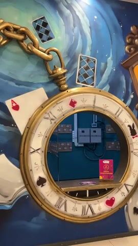 The video pans across a wall mural that appears to be inspired by Alice in Wonderland. A large, ornate pocket watch with Roman numerals dominates the center of the frame, its face serving as a mirror reflecting electrical outlets and a shelf with a small sign. Playing cards, a teacup, and a rose are depicted in the mural, along with a distorted clock. To the right, two couches are visible, one dark gray and the other teal. The camera then moves towards the entrance of a restaurant called "FUN DAY RESTORAN," which features a decorative gold archway and a display of large chess pieces.
