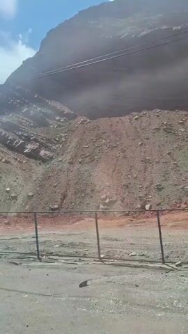 The video shows a large rocky mountain under a clear blue sky with scattered clouds. The mountain's surface is a mix of gray rock layers and reddish-brown earth and loose rock. A road runs along the base of the mountain, separated by a black fence. The camera pans slightly upwards, showing the entirety of the mountain face. There is no visible human activity.