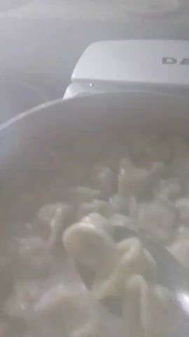 The image shows a close-up, overhead view of a pot filled with dumplings, likely in a boiling liquid. The dumplings are pale and appear to be of a uniform shape. A metal utensil, possibly a spoon or ladle, is partially submerged in the pot, stirring the dumplings. Steam rises from the pot, indicating that the contents are hot.

In the background, a white appliance with the partially visible letters "DA" on it is discernible. This could be a kitchen appliance such as a stand mixer or a food processor. The overall lighting suggests an indoor setting, likely a kitchen, with the light source coming from above or the front.

The scene is focused on the cooking process, suggesting a meal preparation or a cooking demonstration. There are no people visible in the frame. The time of day and weather are not discernible from the image. No specific location cues beyond the domestic setting are apparent. The image is somewhat blurry and lacks sharp detail, but the main subject of the dumplings in the pot is clear. The "DA" text on the appliance is the only visible text. The setting is a domestic kitchen, likely in Berezynka, Ukraine, as per the provided location context.