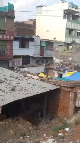 The video displays a view from a moving vehicle, showing a densely populated area with buildings of varying sizes and conditions. Many of the structures appear to be in disrepair. The landscape is littered with trash and debris, suggesting an area of neglect or poverty. There are structures covered with tarps, corrugated metal and brick walls, some of which are painted or contain graffiti. An auto rickshaw is briefly visible. The sky is overcast and the overall atmosphere appears bleak.