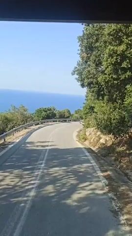 The video shows a scenic drive along a winding road in Palaios Panteleimonas, Greece. The road curves to the right, with a metal guardrail separating it from a steep drop-off. Beyond the guardrail, lush green trees and vegetation cover the hillside. In the distance, the deep blue sea stretches out under a clear, bright sky, suggesting a sunny day with fair weather. The sun casts dappled shadows on the asphalt road, indicating it is likely mid-morning or afternoon. The perspective is from a vehicle moving along the road. There is no visible text in the video.