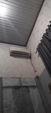 The video shows a close-up view of an air conditioning unit mounted on a wall. The wall is covered in floral wallpaper. A black curtain hangs to the right of the air conditioner. The ceiling appears to be made of corrugated metal. There are wires connected to the air conditioner and a power outlet is visible on the wall below it. The bottom of the frame shows the top of a window with curtains. The video is still and shows no movement or activity.