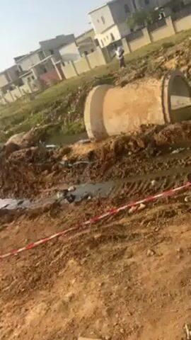 The video shows a muddy construction site on a sunny day. There are puddles of water, and dirt piles are visible. A large concrete pipe sits near a ditch. Red and white striped tape is stretched across part of the area, possibly as a barrier. Buildings are visible in the background, along with some parked vehicles. A few people are seen walking around the site. The camera angle is low, suggesting it's filmed close to the ground.
