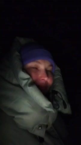 The video shows a person, likely female, in very dark lighting. The individual is bundled in a light-green jacket with a high collar that surrounds their face, and they wear a purple beanie. The person's face is visible, including a mole on their cheek. There are slight head movements. The overall impression is of cold or discomfort. The background is completely black.