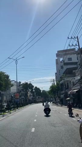 This is a first-person perspective video taken from a vehicle traveling down a paved road. The road has white dashed lines marking the lanes. On either side of the road are buildings, trees, and utility poles with numerous wires. The sky is clear and blue with the sun shining brightly, causing a lens flare. Several motorcycles are visible on the road, moving in the same direction as the camera. One motorcycle is directly ahead, and others are further down the road. Some buildings appear to be residential or commercial, with multiple stories. The overall scene depicts a sunny day in a city or town.
