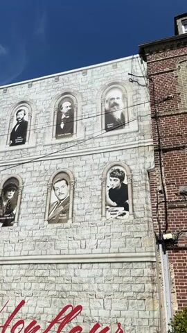 The video shows a mural on the side of a building, depicting portraits of several men and one woman in arched frames. The mural is designed to look like a stone wall with two rows of portraits. The bottom of the mural features large red cursive lettering that reads "J'aime Honfleur", meaning "I love Honfleur". To the right of the mural, a portion of a brick building is visible. The sky is blue with some white clouds. In the foreground, two people are partially visible, one with red hair and another wearing a hat and dark clothing, seemingly pointing towards the mural. A metal fence is also present in the foreground. The overall scene suggests an outdoor public space, likely in a town named Honfleur.