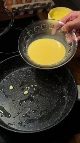 The media shows a close-up of a person's hands preparing to cook an omelet. The scene is set in a kitchen, with a dark-colored non-stick frying pan on a black induction hob. The pan contains a small amount of cooking oil and a few pieces of what appear to be chopped ham or similar ingredients. A glass bowl filled with beaten eggs, also containing some ham pieces, is being held over the frying pan. The eggs are being poured from the bowl into the hot pan, starting to cook and spread out.

In the background, a carton of eggs is partially visible, along with a yellow tub of butter or margarine with "500g" written on it. The lighting suggests it is daytime or the kitchen is well-lit. The overall mood is one of food preparation. There is no visible text directly related to the activity, other than the product information on the butter tub. The context of Słupsk, Poland, indicates the geographical location where this cooking activity is taking place.