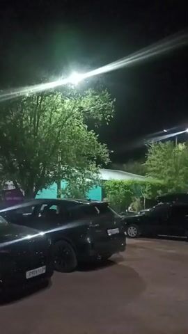 The video shows a nighttime scene in a parking lot, likely in Shubar tubek, Kazakhstan. Several vehicles, primarily SUVs and cars, are parked. The lighting suggests artificial illumination from streetlights, casting long beams across the scene and highlighting the trees and the parking lot surface. The atmosphere is calm and quiet, with no discernible activity or people visible. The presence of cars indicates a place where people might gather or visit, such as a restaurant, event venue, or public area. The weather appears to be clear and dry, typical for an evening in this region. The focus is on the parked vehicles and the surrounding environment under the artificial night lighting.