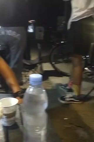 A low-angle, handheld video clip shows a group of people and objects at night. In the foreground, a plastic bottle of water with a blue cap stands on a dark surface, likely a table. To its left, a white paper cup is partially visible. Further left, a drink in a clear cup with a red liquid and a white, crispy topping is on the table. A person's arm, wearing a bracelet, is visible resting on the table near this drink. In the mid-ground, a person wearing black Vans sneakers with colorful socks stands with their back to the camera, wearing a white long-sleeved shirt and black shorts. Their legs are slightly bent. Behind them, bicycles are present, one with a black frame and spoked wheels. To the left of this person, another individual in grey sweatpants with "NEW YORK" printed on the back is partially visible. The background is dark, suggesting an outdoor nighttime setting, with some artificial lighting illuminating parts of the scene. The overall impression is a casual gathering or rest stop.