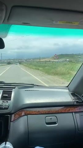 The video shows the interior of a car from the perspective of a passenger. The dashboard and windshield are visible. Outside the windshield, a road stretches into the distance, lined with grassy fields and some buildings. The sky is cloudy. A person's legs are visible in the lower right corner of the frame.