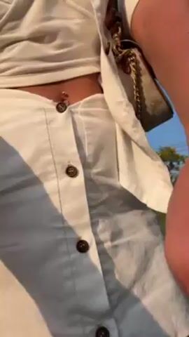 A young woman with wavy brown hair and sunglasses stands in front of a grassy hill against a clear blue sky. She wears a white two-piece outfit consisting of a cropped tank top and a button-down skirt, paired with a matching button-down shirt and a small, patterned purse slung over her shoulder. Her navel piercing is visible. She poses, adjusting her hair and smiling at the camera.