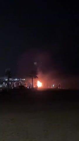 This media file captures a nighttime scene on a beach, likely in Playa Del Sol-Villacana, Spain. The primary focus is a large bonfire crackling brightly, casting an orange glow on the surrounding area. Silhouetted figures of people are gathered around the fire, suggesting a communal event or celebration. Two palm trees stand to the left of the bonfire, adding to the tropical or Mediterranean ambiance. The sky is dark, indicating it is nighttime. The ground appears to be sandy, consistent with a beach setting. While the exact emotions are difficult to ascertain due to the low light and distance, the gathering around a bonfire typically evokes feelings of warmth, community, and festivity. There is no visible text in the media file.