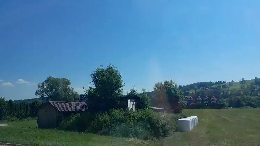 The video captures a scenic view of a rural landscape under a clear blue sky. In the foreground, there is a grassy field with a small, rustic-looking building situated near some trees. To the left, a larger tree stands prominently. As the perspective shifts, a dark-colored car is seen parked near a building with a sign that reads "RESTAURACJA SERKI". Further down the road, a white car drives past, and a large billboard with an image of a horse is visible. The background reveals rolling hills dotted with houses and trees, suggesting a small village or town. Two large cranes are also present in the distance, indicating ongoing construction. The overall atmosphere is peaceful and idyllic, showcasing a typical countryside setting.