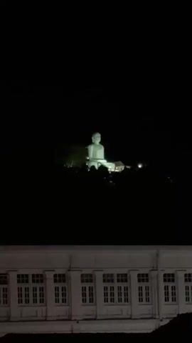 The video presents a nighttime view of a brightly illuminated Buddha statue perched atop a hill, observed from street level. The statue, a light color, stands out against the dark sky. In the foreground, a multi-story building with a white facade and a grid of evenly spaced windows can be seen. As the camera slightly zooms out, additional buildings and passing cars become visible on the street. The scene conveys a sense of urban life juxtaposed with the tranquility of the illuminated Buddha statue.