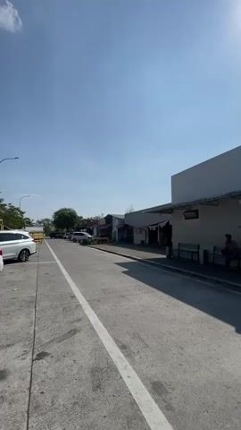 This is a vertical, first-person perspective video of an empty street on a clear, sunny day. The street is paved and has a white line down the center. To the left of the street is a sidewalk and a parking area with a white car. To the right of the street is a building with a white facade and a covered walkway. There are benches outside the building. The sky is blue with no clouds. The camera is slowly panning to the right.