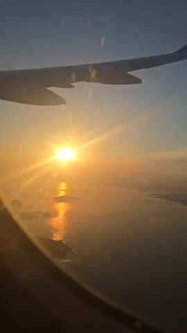 The video captures a serene view from an airplane window during golden hour. A prominent airplane wing occupies the upper left portion of the frame. Below the wing, the sun, a brilliant orb of light, is positioned low on the horizon, casting a radiant glow across the sky and reflecting brightly on the surface of a large body of water. Several landmasses, possibly islands or coastal areas, are visible within the water, appearing dark in silhouette against the sun's reflection. The sky transitions from a deep blue at the top to a warm orange and yellow hue near the horizon, indicative of either sunrise or sunset. The sun's intense rays create a starburst effect. The scene evokes a tranquil and picturesque moment high above the earth.